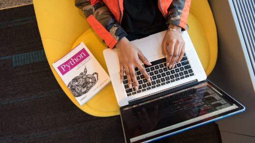 Person coding on a laptop beside a Python programming book and a tablet displaying code.