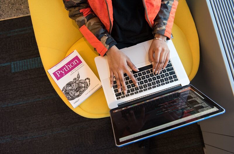 Person coding on a laptop beside a Python programming book and a tablet displaying code.
